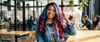 Young woman with blue and pink ombre hair wearing denim shirt in modern cafe setting, lifestyle portrait for fashion and beauty content.