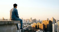 Young man sitting on rooftop edge overlooking urban cityscape at golden hour sunset, contemplating future opportunities and life decisions.