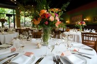 Elegant wedding reception table setting with colorful floral centerpiece featuring orange and pink blooms, white linens, formal place settings and ambient lighting.