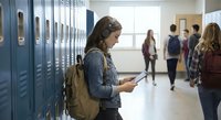 Teenage girl using smartphone in school hallway with lockers while students walk in background during break time.