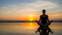 Woman meditating in lotus pose on beach at sunset with golden sky reflection on wet sand creating peaceful zen moment for wellness and mindfulness practice.