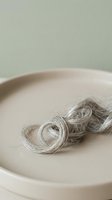 Twisted gray hair strands arranged in spiral loops on a neutral ceramic plate against a soft sage background, suitable for minimalist styling, wellness, and beauty editorial use.