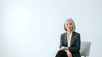 Senior Asian woman with gray hair wearing dark blazer and white blouse, seated in modern chair against white background. Professional portrait for corporate, business, leadership, and executive concepts.
