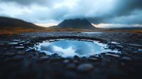 Dramatic mountain landscape reflected in small water puddle on rocky ground during moody sunset, with dark stormy clouds and atmospheric lighting creating mirror effect.