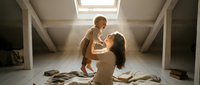 Young Caucasian mother lifting laughing baby in sunlit attic room, sharing a joyful tender moment surrounded by warm natural light.