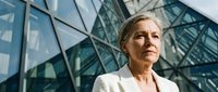 Mature woman with gray hair wearing white blazer standing against modern glass building facade with geometric architecture, professional business portrait.