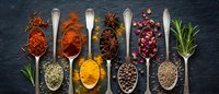 Colorful spices and herbs arranged in wooden spoons on dark slate background. Culinary ingredients including turmeric, paprika, star anise, peppercorns and rosemary.