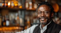 Mature African American man with glasses smiling in bar or restaurant with blurred bottles in background, warm ambient lighting creates inviting atmosphere.