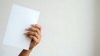 Close-up of a hand with medium skin tone holding a blank white paper sheet against a neutral background, suitable for mockups and presentations.