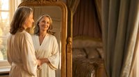 Mature blonde woman in white bathrobe looking at reflection in ornate golden mirror in luxury bedroom interior for wellness and self-care concepts.
