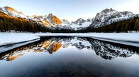 Snowy mountain peaks reflect in calm alpine lake at sunrise, dramatic winter landscape with golden light on rocky summits and pine forest shoreline.