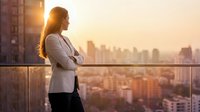 Professional businesswoman in white blazer standing on balcony overlooking city skyline at sunset, contemplating future success and career growth opportunities.