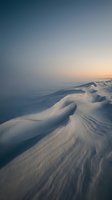 Windswept snow drifts create flowing patterns across winter landscape during golden hour twilight with soft pastel sky gradient for seasonal backgrounds.