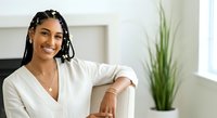 Young woman with dark braided hair and medium brown skin tone wearing white casual shirt, smiling at camera in bright modern interior with potted plant, suitable for lifestyle and wellness content.