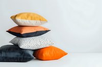 Stack of modern decorative pillows in orange, black, and white with geometric patterns and textures against light wall, showcasing contemporary home decor style.