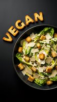 Vegan Caesar salad with romaine lettuce, croutons, capers, and shaved cheese in dark bowl, with word VEGAN spelled by croutons on black background.