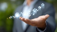 Professional businessman in gray suit holding glowing digital chart visualization with rising trend bars and sparkles floating above palm against blurred background.