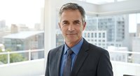 Confident mature Caucasian businessman in dark suit and tie standing in modern office with city view background for corporate and professional use.