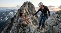 Two climbers helping each other on rocky mountain peak during golden hour sunset with dramatic alpine landscape background for adventure sports content.