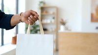 Person with dark skin tone holding white shopping bag with handles in modern retail environment, representing consumer purchase and retail experience.