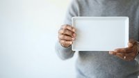 Man in gray sweater holding blank white tablet for mockup, advertising, or promotional content display against neutral background.