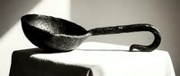 Ancient bronze ladle with ornate handle displayed on white fabric surface in dramatic lighting for historical archaeology museum exhibition.
