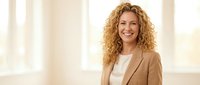 Professional businesswoman with curly blonde hair smiling confidently in beige blazer and white top, standing in bright modern office environment.
