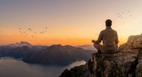 Man meditating on mountain cliff at sunset overlooking lake with flying birds in peaceful landscape for wellness and mindfulness concepts.