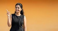 Young Indian businesswoman pointing upward with finger against orange background, professional attire, confident expression for presentations.