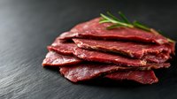 Fresh sliced rare roast beef with rosemary garnish arranged on dark slate background, raw meat texture visible, high protein food photography.