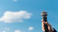 Hand holding professional microphone against bright blue sky with white fluffy clouds, representing freedom of speech, communication and public speaking.