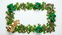 Flat lay frame with lucky charms, green hats, horseshoes, coins and clover leaves on white background for St. Patrick's Day celebrations and festive designs.