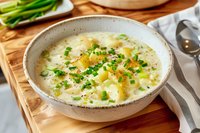 Creamy potato leek soup garnished with fresh chives and black pepper in rustic ceramic bowl on wooden board, comfort food styling for restaurant menu.