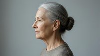 Senior woman with gray hair in profile, wearing neutral beige sweater against gray background. Mature adult portrait for healthcare, wellness, aging, and lifestyle editorial use.