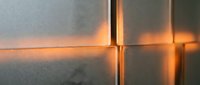 Warm golden light glowing through seams of matte gray tiles, creating dramatic contrast and depth on a modern textured wall surface.