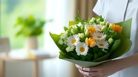 Fresh spring bouquet with white daisies, orange tulips and delicate baby's breath wrapped in green paper, held by hands against blurred interior background.