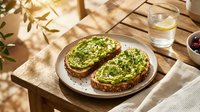 Healthy avocado toast on rustic bread served on wooden table with natural sunlight and shadows creating warm morning breakfast atmosphere.