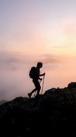 Silhouette of female hiker with trekking poles ascending rocky mountain peak at sunrise above clouds, adventure and outdoor fitness concept.