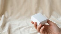 Unboxing white product box in hand against neutral beige fabric background for packaging and branding mockups.