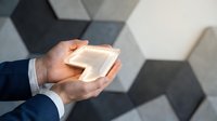 Businessman in dark suit holding glowing white cube against geometric hexagonal wall background, representing innovation, technology, and modern business solutions.