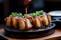 Pouring sweet soy sauce over pan-fried dumplings garnished with fresh green onions on black plate, close up view of Asian cuisine with dramatic lighting.