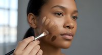 Young African American woman applying foundation makeup with brush testing different skin tone shades on face for perfect color match in natural light.