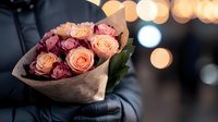 Fresh pink and peach roses wrapped in kraft paper, held by person wearing dark coat against blurred bokeh lights background, romantic gift concept.