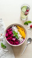 Overhead view of a vibrant acai bowl topped with diced beetroot, mango, coconut flakes, and mint leaves, served with a refreshing cucumber and berry infused water on a light wooden surface.