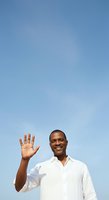Man with dark skin tone wearing white button-up shirt waving hand in greeting against clear blue sky, conveying friendliness and approachability for business, communication, and lifestyle concepts.