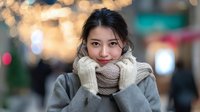 Young Asian woman wearing cozy knit scarf and mittens smiling warmly in winter city street with bokeh lights background for seasonal lifestyle content.
