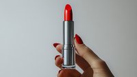 Red lipstick in silver tube held by female hand with matching manicure on neutral background for beauty and cosmetics advertising campaigns.