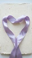 Purple satin ribbon arranged in heart shape on textured white paper background for romantic wedding invitation design and craft projects.