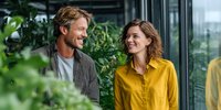 Cheerful Caucasian couple enjoying conversation in modern office space with lush green plants, creating positive work environment.