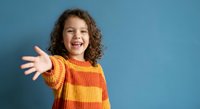 Happy young girl with curly hair wearing striped sweater reaching out with open arms on blue background expressing joy and excitement for childhood concepts.
